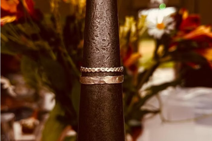 Two thin silver bands, one with a textured hammered finish and the other with a wavy pattern, displayed on a dark metal ring holder in a warm, blurred event setting with flowers in the background.