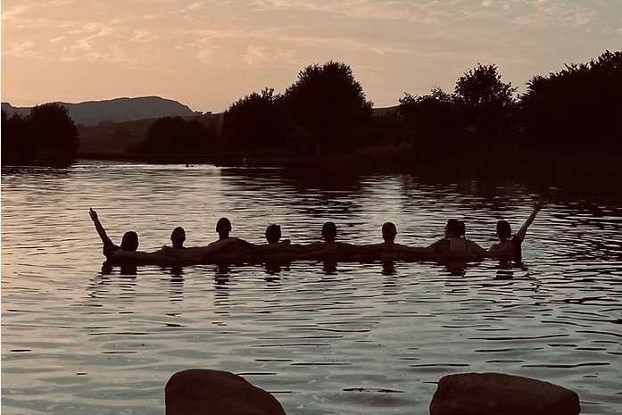 Silhouetted figures sitting in a long canoe on a lake at sunset, with trees, distant hills, and two large rocks in the foreground.